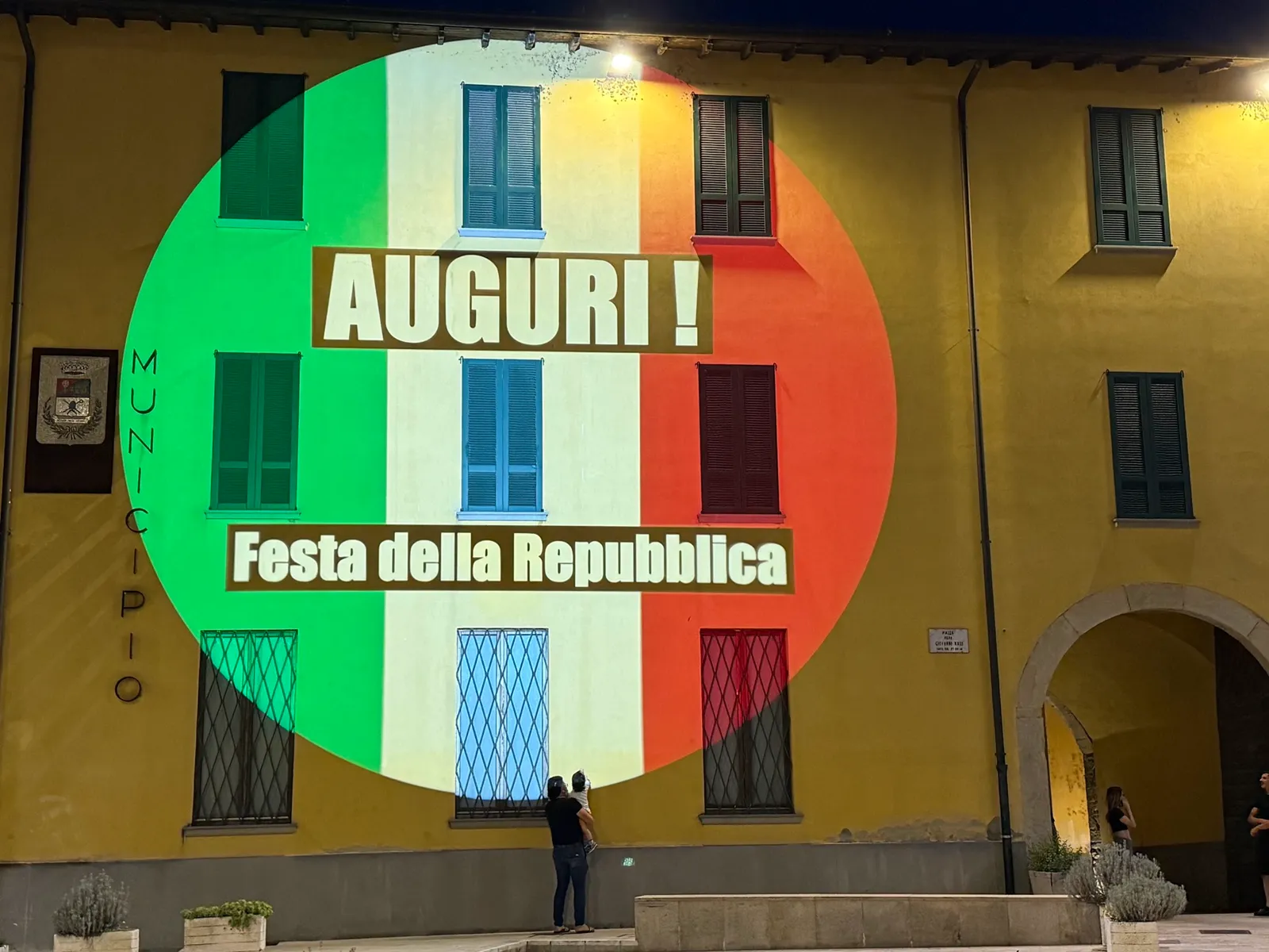 Tricolore proiettato in un cerchio sulla facciata gialla del municipio di Ghisalba, con le scritte Auguri e Festa della Repubblica: 2 giugno 2025