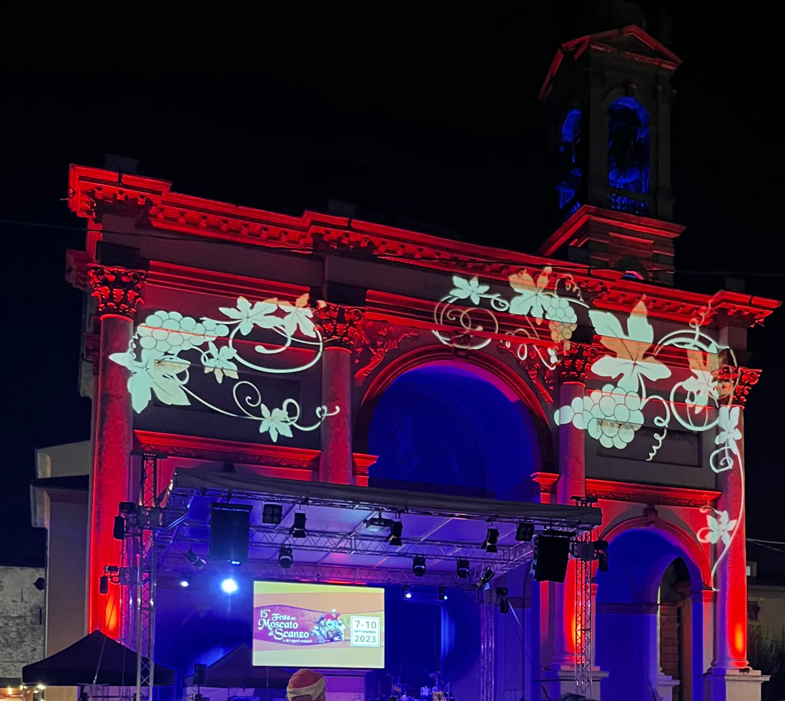 Tralci di vite con grappoli proiettati sulla facciata della chiesa illuminata di rosso e blu, con il palco davanti, alla Festa del Moscato di Scanzo di Rosciate, 2023